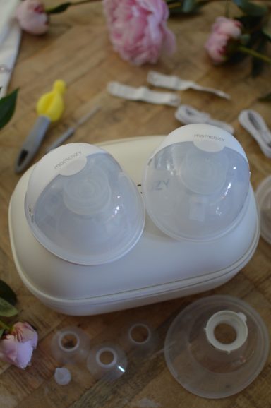 That Mama Club x Momcozy A Momcozy wearable breast pump sits on a wooden table surrounded with the many accessories that it comes with.