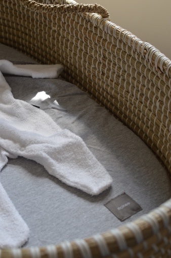 That Mama Club x MoKee A close up of a baby Moses basket. The basket is made from woven seagrass in a natural colour, with grey fitted sheet and white baby onesie.