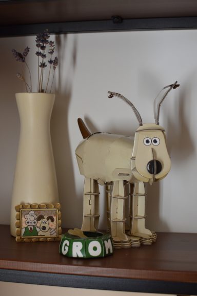 That Mama Club x BYO Kits A cardboard model of Grommet from Wallace and Grommet sits on a wooden shelf next to a cream vase with dried lavender flowers.